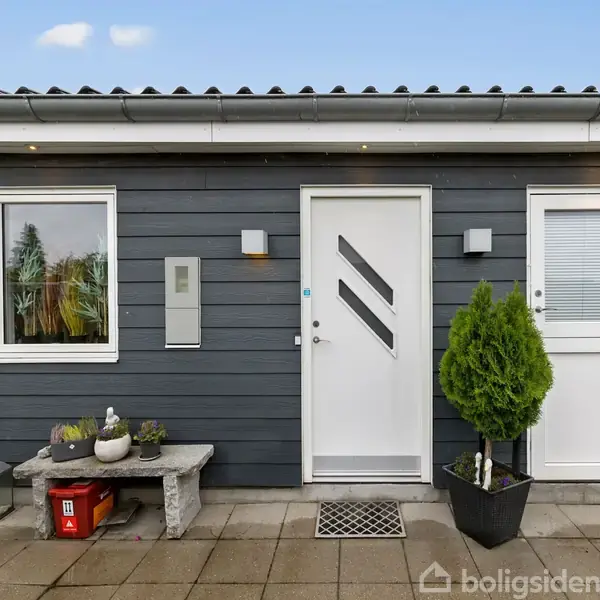 En moderne bygning med grå facade; to vinduer og en hvid dør. En lille grøn plante står i en sort potte ved siden af døren på en flisebelagt terrasse.