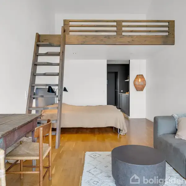 En stue med en grå sofa, et rundt bord og et skrivebord med stole. En trappe fører op til en hems. Rummet er lyst med billeder på væggen og en seng i baggrunden.