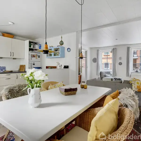 En hyggelig stue med spisebord fyldt med blomster i vase, omgivet af en lys sofa og køkkenområde. Store vinduer lader lys komme ind, med udsigt til en bybygning.