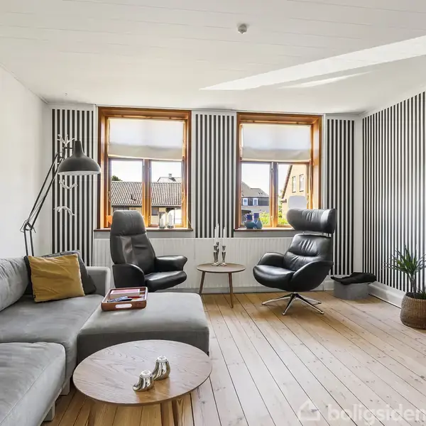Gråt sofaarrangement med puder og sofabord med kaffekopper. To lænestole er placeret mod stribede vægge med tre vinduer. Trægulve og skuffer, tv til højre, plante ved siden af.
