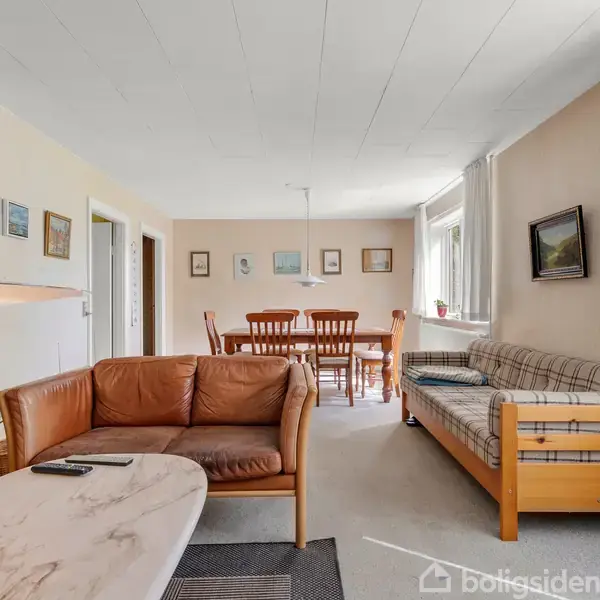 Brunt lædermøblement med sofabord i stue; spisebord med træstole i baggrunden. Lyse vægge dekoreret med billeder, store vinduer med gardiner til højre.