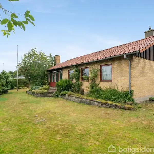 Et murstenshus med rødt tegltag står stille i en grøn have. Buske nær husets facade; græsplæne omgiver området. Klar himmel i baggrunden.