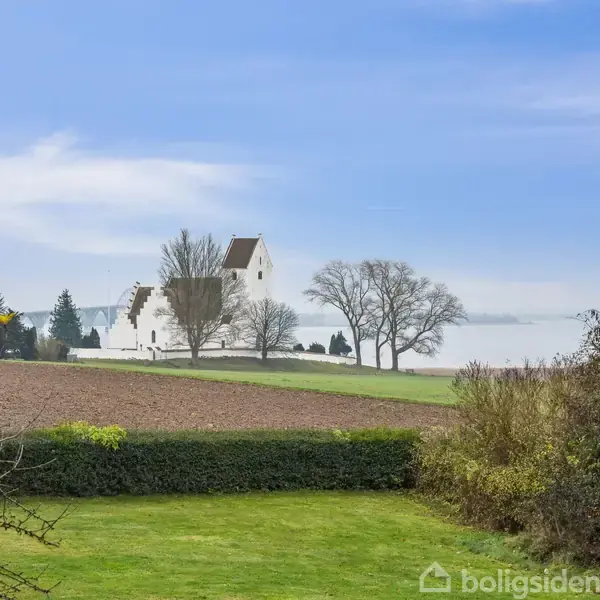 En hvid kirke står på en bakke med træer omkranset af marker og grønt græs. Klare himmelstrøg danner baggrunden i en landlig scene.