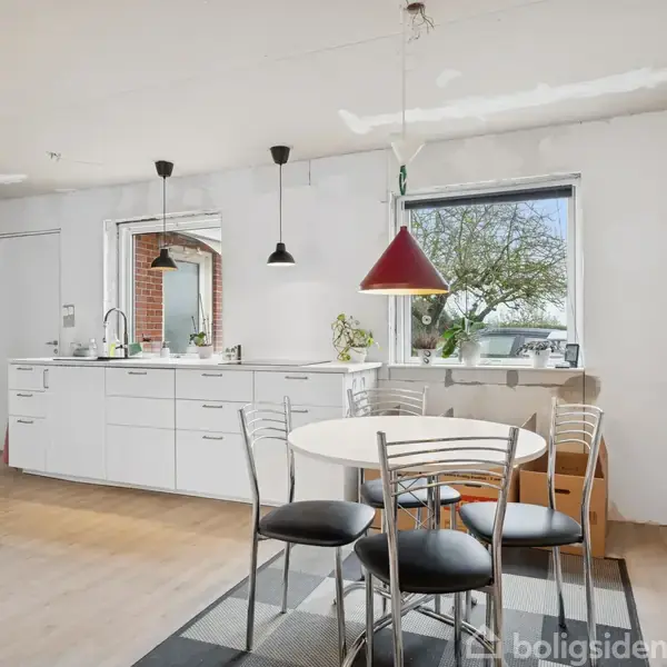 Et lyst køkken har et rundt bord med stole og moderne pendellamper. Køkkenskabene er hvide, og store vinduer giver udsigt til en have med et træ.