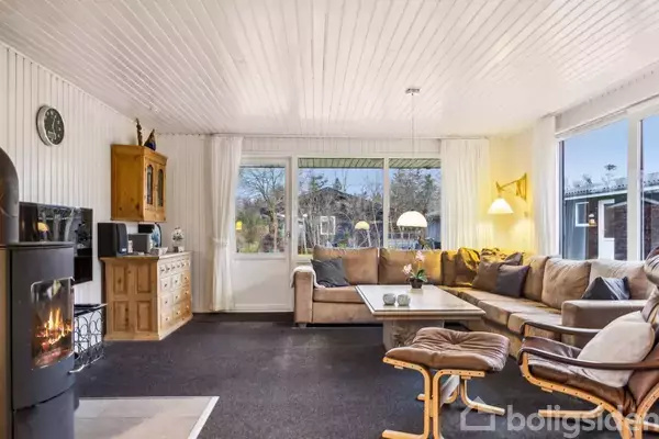 En stue med en stor beige hjørnesofa omkring et sofabord. En pejs til venstre og store vinduer giver udsigt til haven. Lyse vægge og træmøbler.