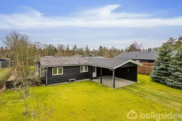 Et sort træhus med carport står på en velplejet græsplæne. Omgivet af buske og træer, ligger det i et fredeligt beboelsesområde under en blå, let overskyet himmel.