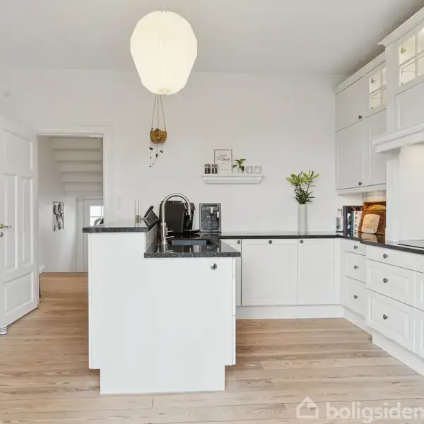 Køkken med hvide skabe og sorte bordplader. En central ø med vask står i midten. Lyse trægulve og en enkelt hængende lampe. Blomster og dekorationer tilføjer charme.