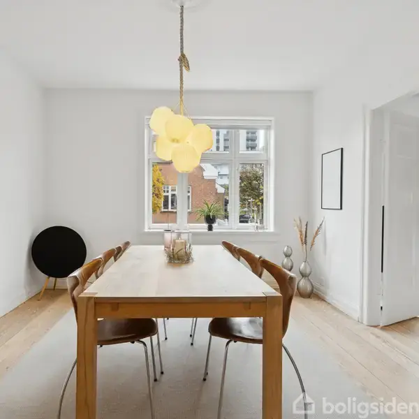 Lang spisebord med seks stole står i et lyst rum. En moderne lyslampe hænger over bordet. Store vinduer og farverigt maleri dekorerer væggene.