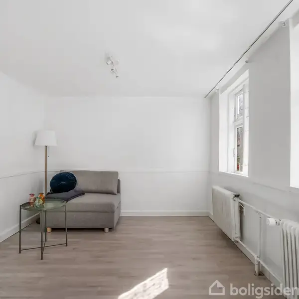 Sofa står i et hjørne med en pude på, ved siden af et glasbord med drikkevarer. Rummet er lyst med hvide vægge, trægulv og vinduer mod en murstensbygning.