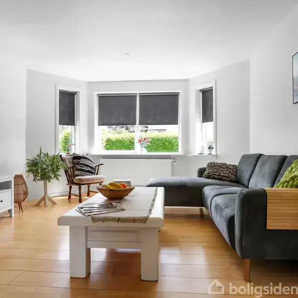 Moderne stue med grå sofa og sofabord i træ; fjernsyn på hvidt mediebord til venstre. Store vinduer med sorte rullegardiner og udsigt til grøn have.