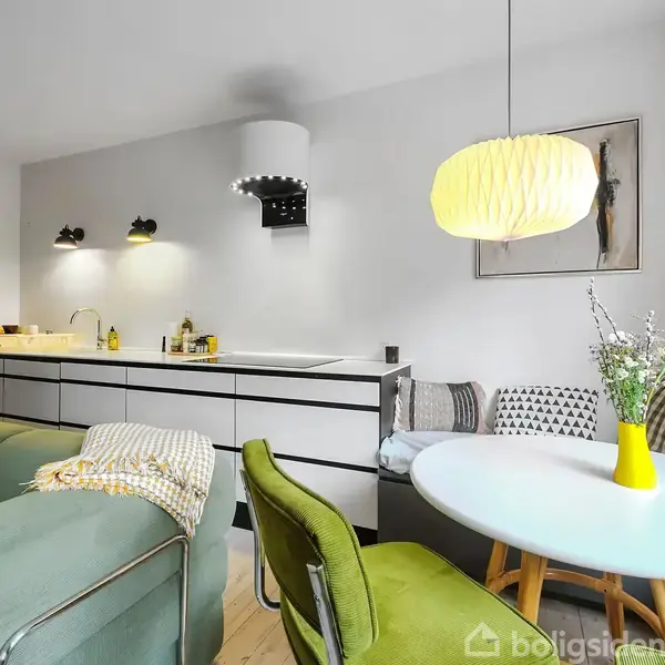 Et moderne køkken med hvide skabe. En grøn sofa står ved siden af et rundt bord med en gul vase. En dekorativ lampe hænger over bordet.