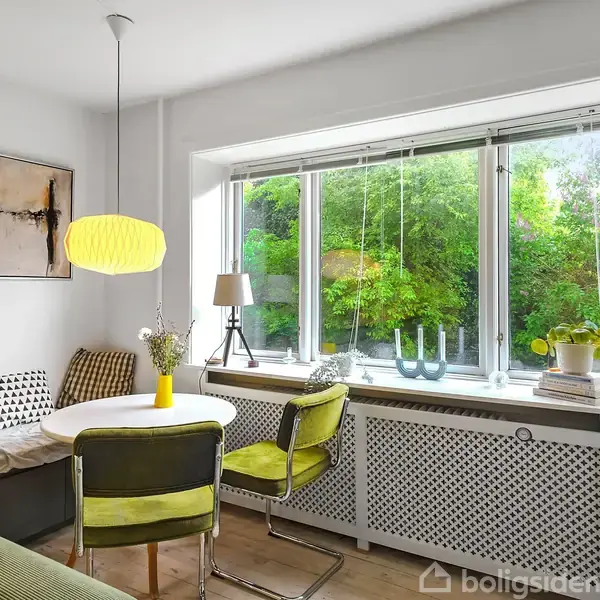 Et lyst køkken med et rundt spisebord omgivet af stole og en bænk. En stor vindueskarm med udsigt til træer indeholder planter og lamper.