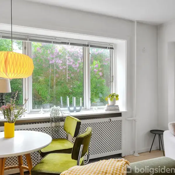 En lys stue indeholder et rundt bord med blomster og en gul lampe hængende over. Grønne stole står i rummet, der har store vinduer med udsigt til en have.