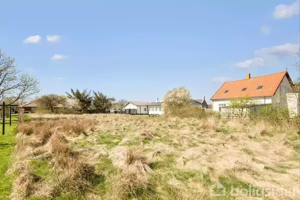 En åben mark med høj græs, omkranset af et hus med rødt tag til højre. I baggrunden ses flere huse under en klar blå himmel.