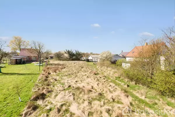 En grund med vildtvoksende græs strækker sig mellem to huse i en landlig omgivelser, omgivet af træer og en klar blå himmel i baggrunden.