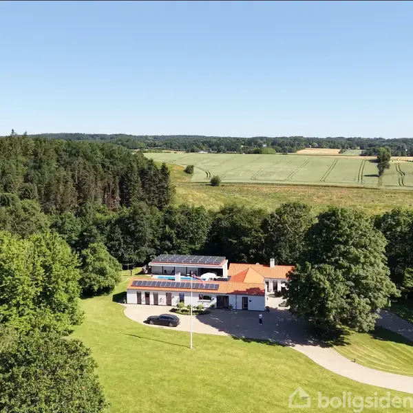 Et hus med solpaneler på taget står i en lysning omgivet af træer. Der er marker i baggrunden, og himlen er klar og blå.