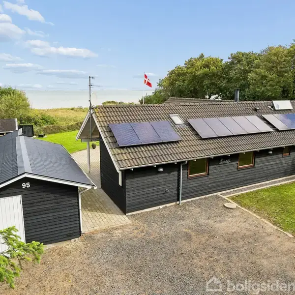 Sort hus med solpaneler på taget, beliggende i grønne omgivelser. Dansk flag vajer i baggrunden. Lille skur med nummer 95 står til venstre, omgivet af træer og græsplæne.