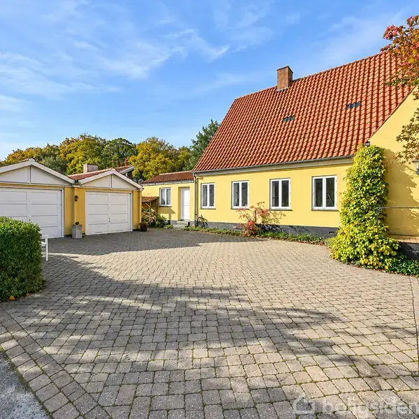 Gult hus med rødt tegltag står stille ved stenet indkørsel, omgivet af grønne buske og træer under en blå himmel. To hvidmalede garager er til venstre.