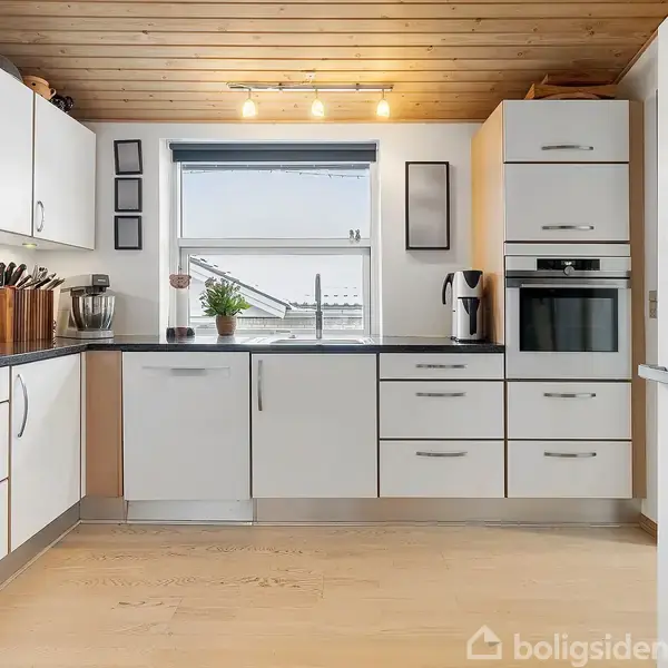 Køkken med hvide skabe, stående og indbyggede apparater. Vinduet centralt sikrer lysindfald. Trægulv og -loft skaber en varm atmosfære. En plante står på bordpladen ved vasken.