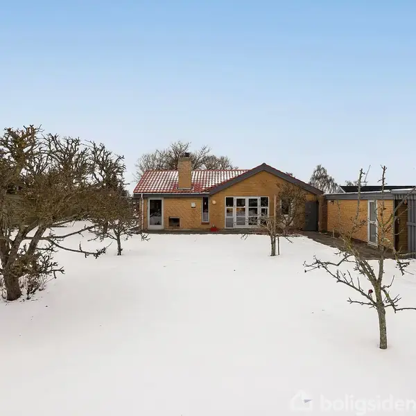 Et gult hus med rødt tag står i en snedækket have med få nøgne træer. Til højre ses en grå bygning eller skur. Himlen er klar og blå.