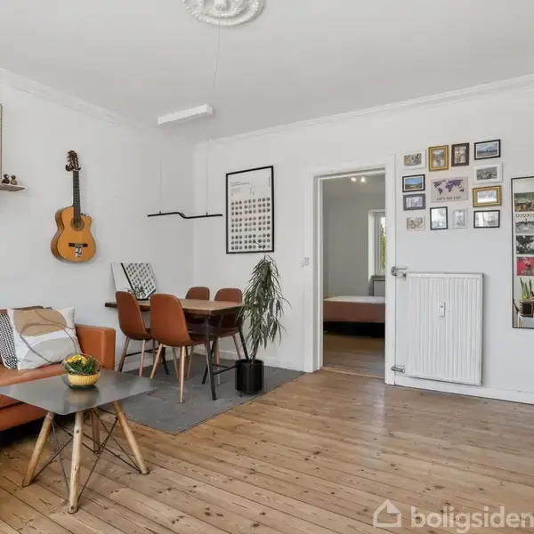 En stue med en brun hjørnesofa står ved siden af et lille sofabord. En guitar hænger på væggen overfor et spisebord med fire stole. Væggene er dekoreret med billeder.