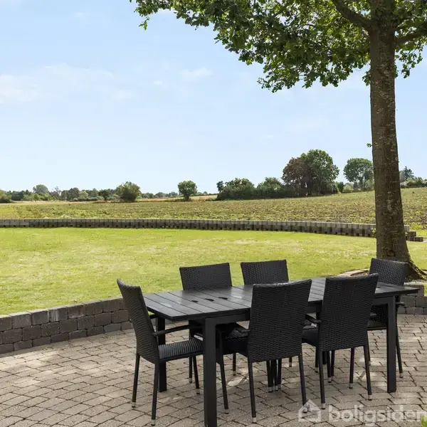 Havebord med seks stole står på en brolagt terrasse. Et stort grønt græsareal omgiver terrassen, med en træ omkranset af marklandskab og en murstensvæg i baggrunden.