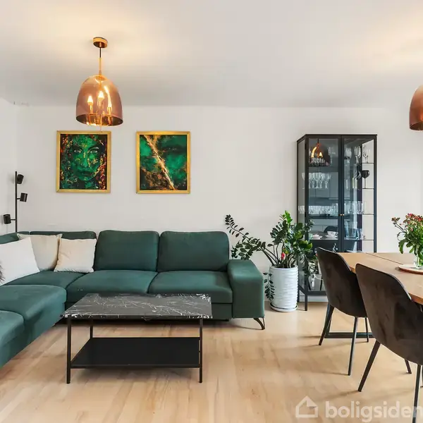 Grøn sofa står i moderne stue med marmorbord foran, tre malerier hænger på væggen. Ved siden af er en sort vitrineskab, og spisebord med stole står til højre.