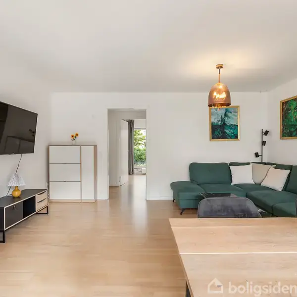 Grønt hjørnesofa står i en lys stue med trægulv, flankeret af tre malerier. Et TV er vægmonteret til venstre, og en rund lampe hænger fra loftet.