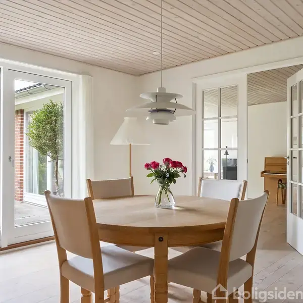 Et rundt spisebord med fire stole står i en lys spisestue. En vase med blomster pryder midten. Store glasdøre åbner ud til en terrasse.