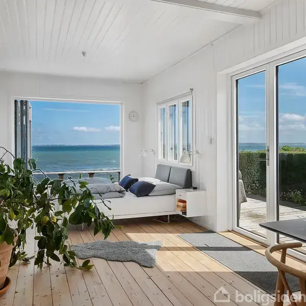 Et lyst værelse med en sofa og skrivebord vender ud mod en terrasse med havudsigt. Et grønt stueplante står ved siden af. Væggene er hvide med træpaneler.