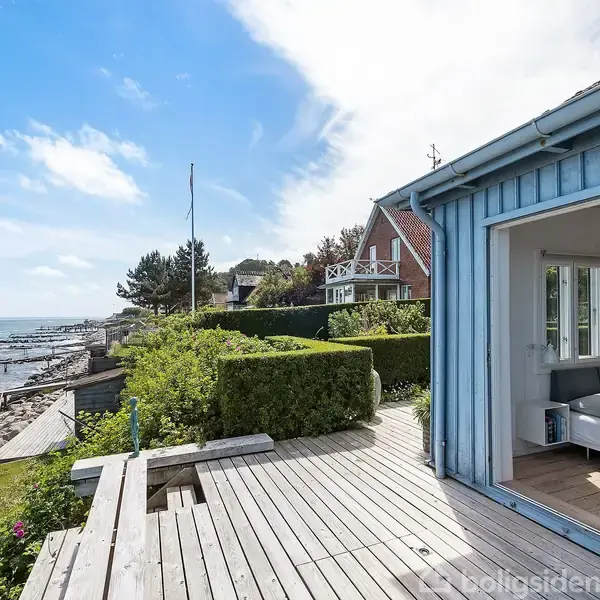 Blå strandhytte med åbne døre, afslører en soveplads der vender ud mod en træterrasse. Kysten med bølger og klipper strækker sig ved siden af, under en klar himmel.