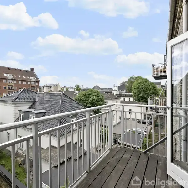 En åben altan med metalrækværk strækker sig ud fra en bygning, omgivet af byens tage og træer under en klar blå himmel med skyer.