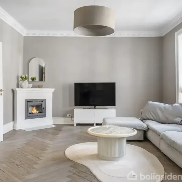 Stue med grå vægge, hvor en grå sofa står ved et vindue. Et rundt sofabord er foran. En fladskærms-TV står ved en pejs med spejl over, omgivet af potteplanter.