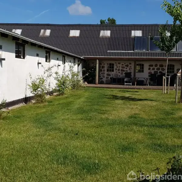 Et hvidt, lavt stuehus med sort tag ligger ved siden af en græsplæne. Husets facade har både mursten og pudsede elementer. Flere vinduer og en solrig, klar himmel.