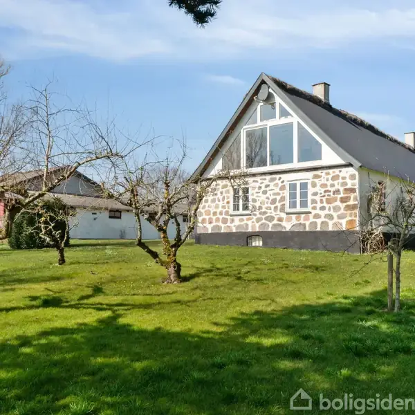Et stuehus med stengavl og store vinduer står på en grøn græsplæne. Omkring huset er der spredte træer, og himlen er blå med lette skyer.