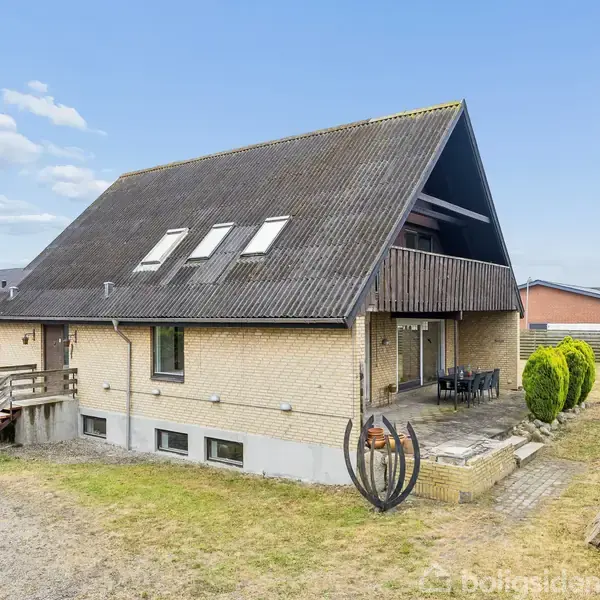 Et to-etagers hus med skråt tag og flere ovenlysvinduer, omgivet af græsplæne og buske. Der er en overdækket terrasse med spisebordsmøbler ved siden af indgangen.