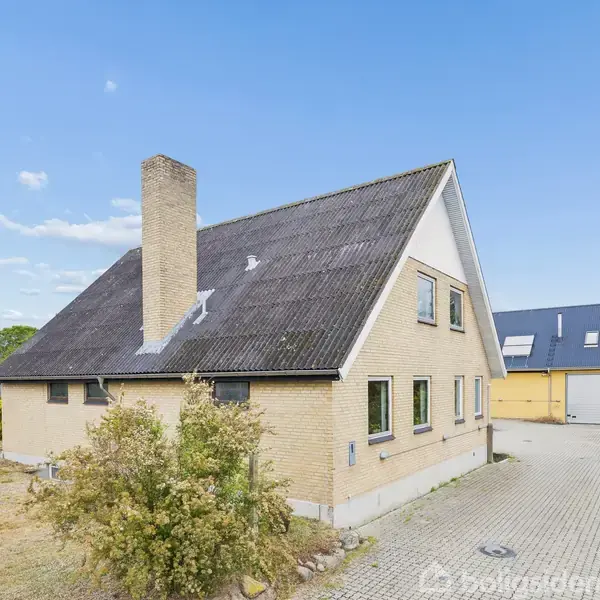 En murstenshus med mørkt skråt tag og skorsten står ved siden af en flisebelagt indkørsel, omgivet af en græsplæne og træer under en blå himmel.