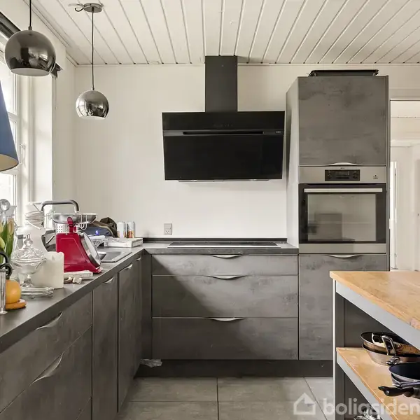 Et moderne, gråt køkken har en indbygget ovn og emhætte. Køkkenbordet er fyldt med køkkenudstyr, herunder en lampe og blender. Det er lyst op af naturligt lys fra vinduerne.