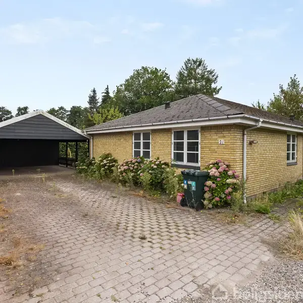 Et gult, muret hus står ved en flisebelagt indkørsel omgivet af buske og træer. Ved siden af er en carport. Nummeret "92" er synligt ved indgangspartiet.
