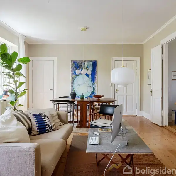 En lys stue med en beige sofa dækket af puder foran et sofabord med en computer. I baggrunden ses et spisebord med stole, kunst på væggen og en stor grøn plante ved vinduet. Der er en pejs i højre side.