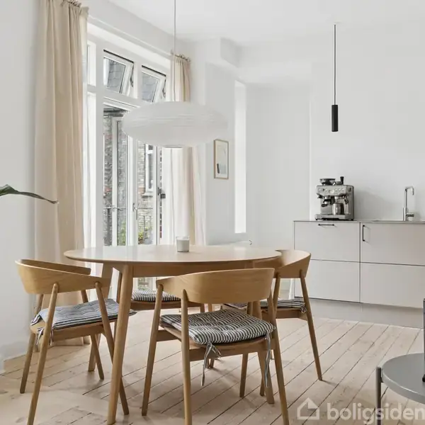 Et rundt træbord med fire stole står i en lys stue. Store vinduer med beige gardiner lader naturligt lys ind. En plante står ved siden af bordet, og en hvid pendellampe hænger over bordet. I baggrunden ses et moderne køkken med grå skabslåger og en kaffemaskine.