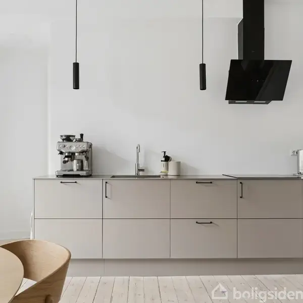 Køkkenet har en minimalistisk, moderne stil med grå skabe, en sort emhætte, og en kaffemaskine. Der hænger to sorte pendellamper fra loftet. Rummet er lyst med lyse trægulve.