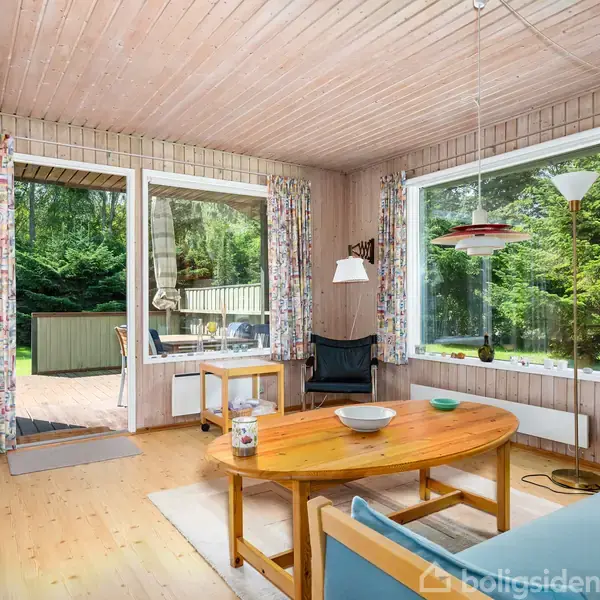 Stue med træmøbler; en sofa og rundt sofabord. Store vinduer og terrassedør giver udsigt til have med træer. Gardiner med mønster, væglampe, og en rød lampe hænger fra loftet.