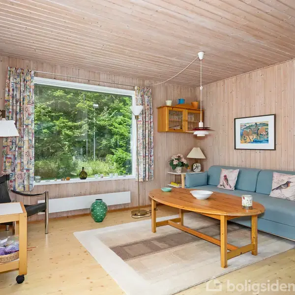 Sofa står i stuehjørne ved vindue med udsigt til grønne træer. Kaffebord foran sofa, lampe og maleri på væggen. Rullevogn ved siden af sofaen.