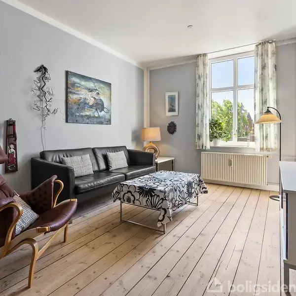 Stue indeholder en sort sofa og brun lænestol omkring et sofabord med blomsterdug. Væggene har kunst og en stålampe. Store vinduer giver naturligt lys.