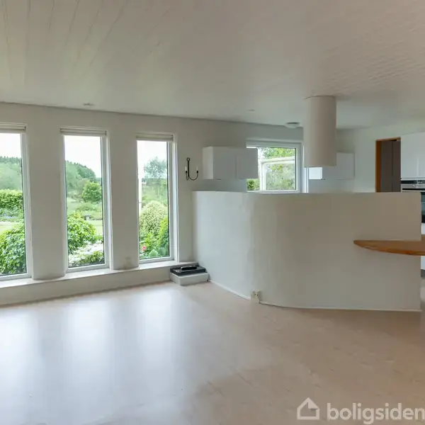 Stue med hvide vægge og gulv, store vinduer viser grøn have. Køkkenområde med hvidt møblement og indbygget ovn til højre. Afrundet køkkenbar.