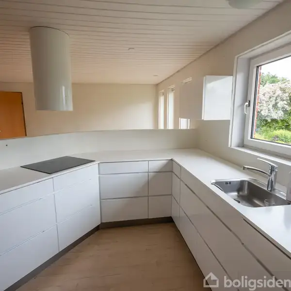 Et moderne køkken med hvide skabe står ubenyttet. En kogeplade er indbygget i bordpladen. Vindue mod en have lader naturligt lys trænge ind, mens en sæbebeholder står ved vasken.