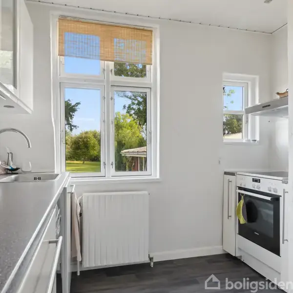 Et lyst køkken med hvide skabe og ovn til højre. En vask er placeret under et vindue med udsigt til en grøn have. Gulvet er mørkt.