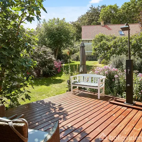 En hvid bænk står stille på en træterrasse. Terrassen er omgivet af en frodig have med blomster, buske og træer, og et hus med rødt tag er synligt i baggrunden.