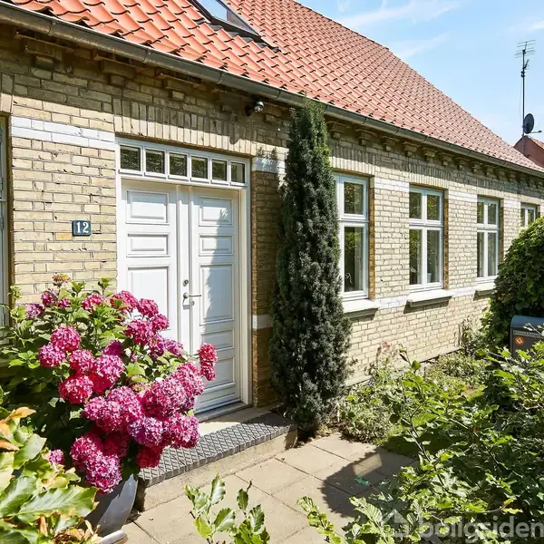Et murstenshus med rødt tegltag står i en have med blomstrende buske. Huset har hvide vinduer og en hoveddør. En lille plakette viser nummeret 12.